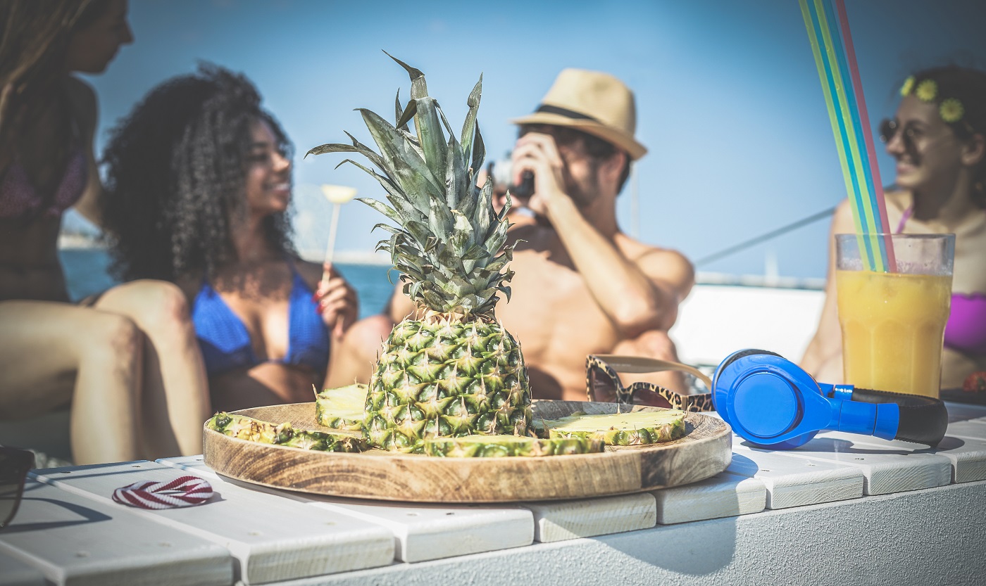 Happy friends having fun and eating pineapple fruit at boat party - Young people taking photos in caribbean sea tour - Tropical, travel and summer vacation concept - Focus on fruit - Soft vignette